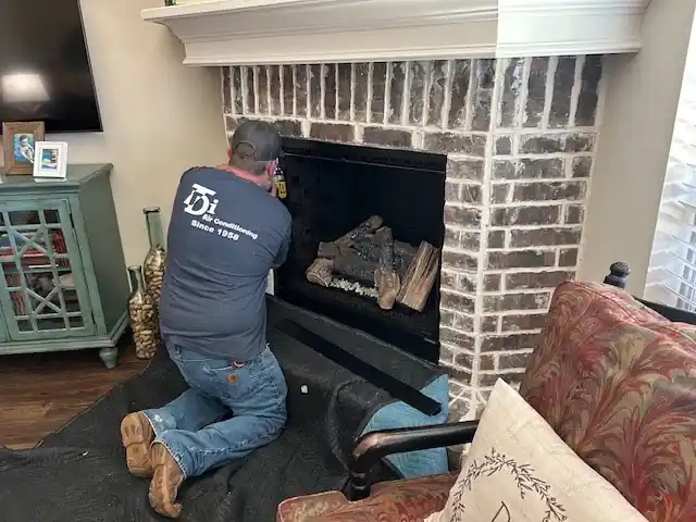 A TDi technician in a Since 1958 shirt, seen from behind, installing gas logs in a brick fireplace